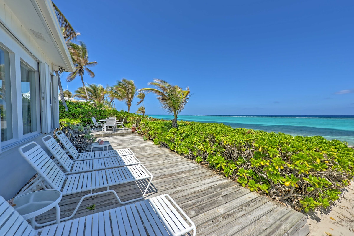 A beachfront patio is shown with several white lounge chairs arranged on a wooden deck. Lush greenery borders the space, leading to the calm turquoise waters of the Caribbean Sea. A clear blue sky complements the serene coastal setting.