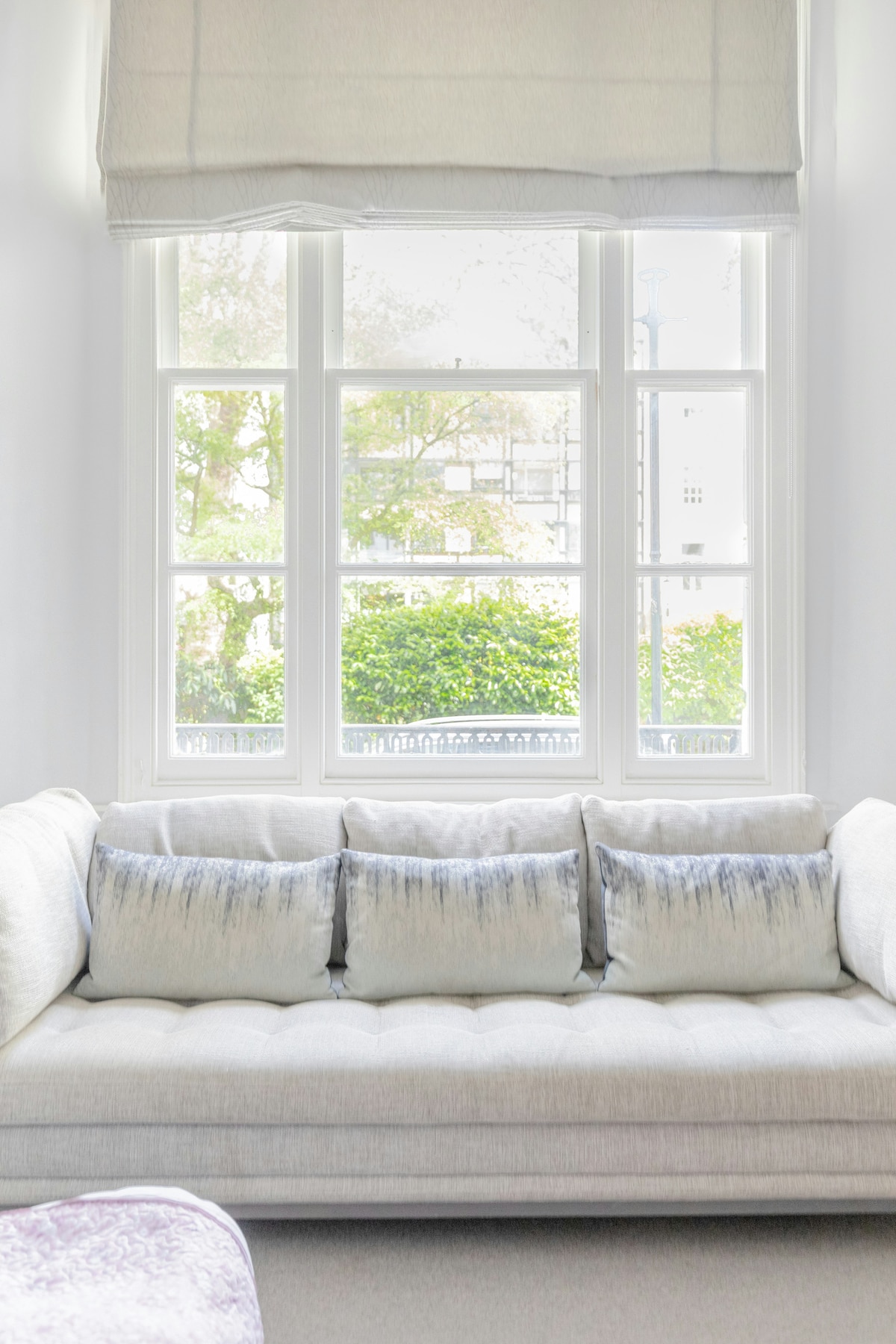 A light-filled living area features a neutral-toned sofa adorned with plush, textured cushions. Large windows provide a view of the lush greenery outside, enhancing the room's airy feel.