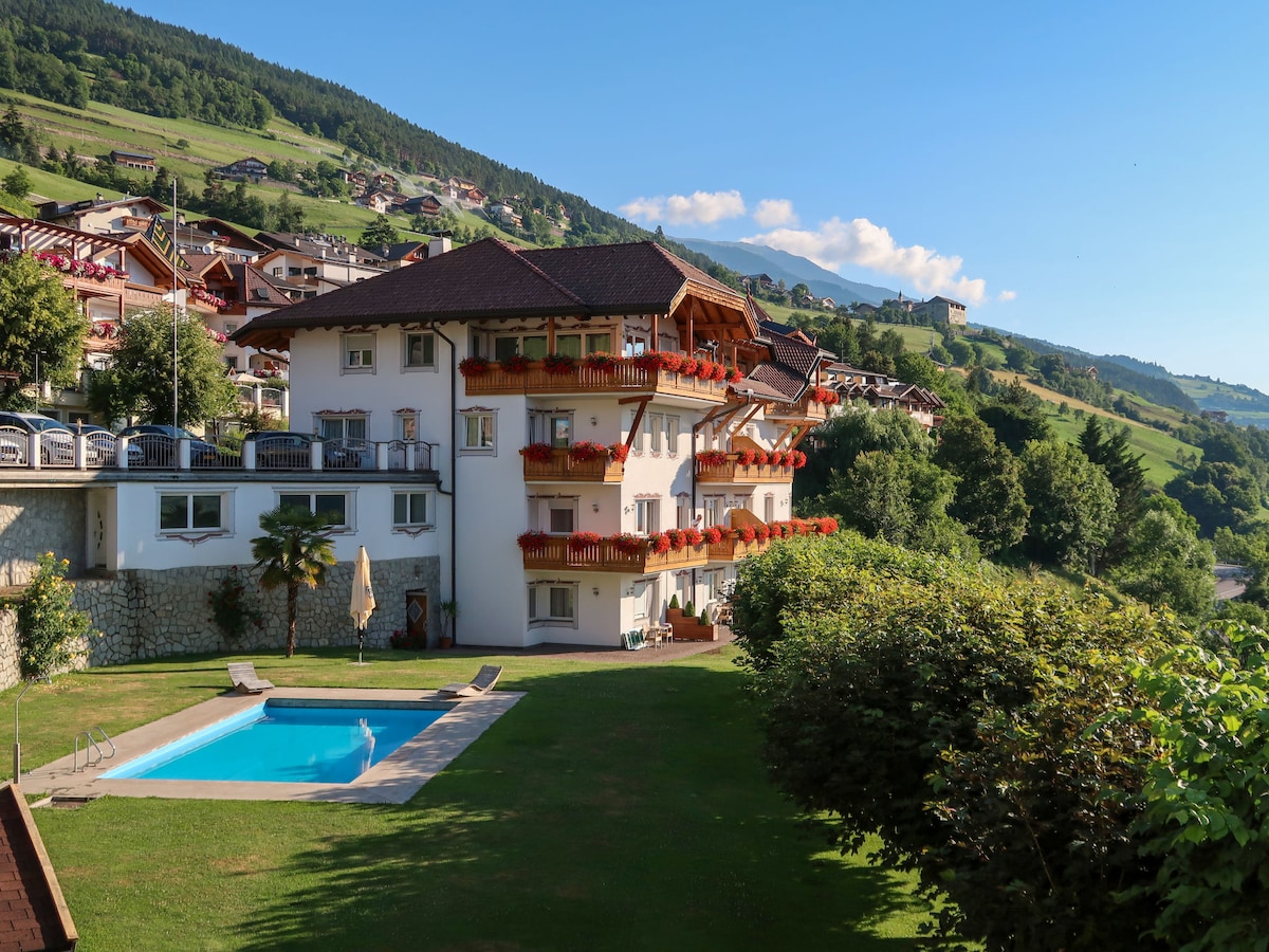 The building displays a charming alpine architecture with wooden balconies adorned with flowers. A well-maintained lawn surrounds a small swimming pool. The hillside landscape in the background features a mix of greenery and residential buildings under a clear blue sky.