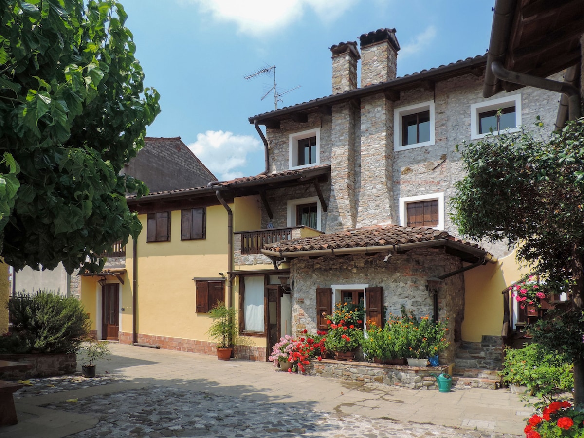 The exterior of the terraced house showcases a blend of rustic stone and warm yellow walls. Lush greenery and vibrant flowers are seen adorning the courtyard, adding color to the charming entryway. A balcony above offers additional outdoor space, while a pathway leads through the courtyard.