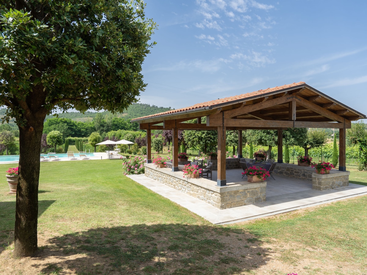 An outdoor terrace is surrounded by flowering plants, providing a shaded area with rustic wooden beams. The adjacent green lawn leads to a swimming pool, complemented by additional sun loungers and umbrellas in a serene setting.