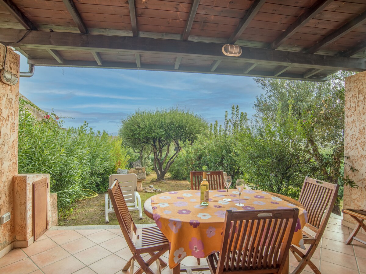 An outdoor terrace features a round dining table with a colorful tablecloth set for four. Wooden chairs surround the table, overlooking a lush garden filled with greenery. A glimpse of the sky can be seen through the terrace's roof structure.