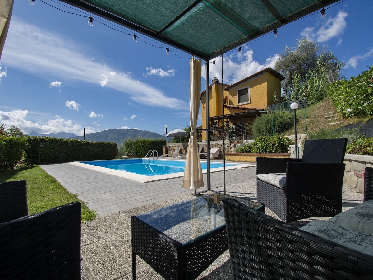 The outdoor area features a swimming pool surrounded by a paved patio, with lounge furniture under a shaded gazebo. A house with rustic architecture is visible in the background, set against a backdrop of hills and clear blue sky.