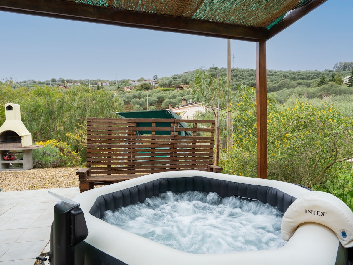 A hot tub is positioned on a patio beneath a shaded structure, providing views of the surrounding countryside. A barbecue area is nearby, and a wooden bench sits adjacent to the hot tub, inviting relaxation in a serene outdoor setting.