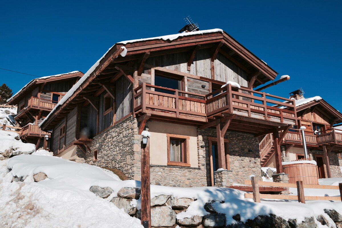 A charming mountain chalet is displayed, featuring a combination of wood and stone materials. Multiple balconies offer a view, while a warm wooden hot tub is visible in the courtyard area. Snow blankets the ground, enhancing the alpine atmosphere.