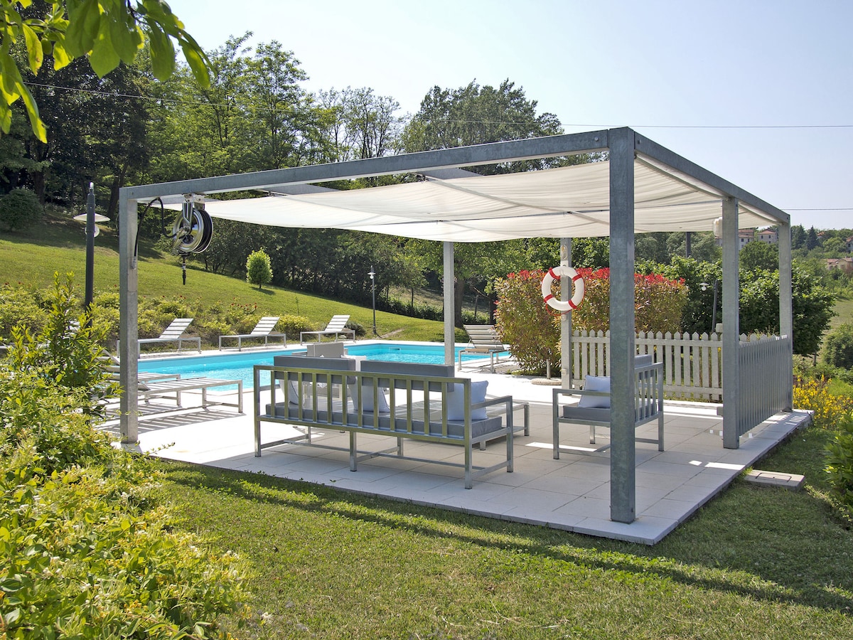 A shaded outdoor seating area is featured under a canopy, accompanied by a pool visible in the background. The space is furnished with a gray sofa set and is surrounded by well-maintained greenery. The area offers a relaxing view of the swimming pool and garden.