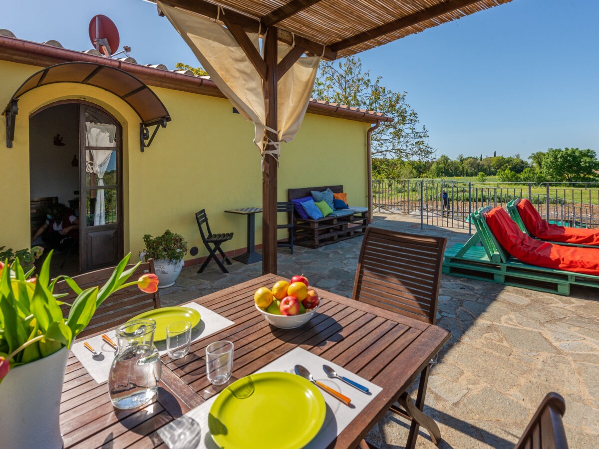 An outdoor dining area with a wooden table is set with plates and glasses, featuring a bowl of apples as a centerpiece. Colorful lounge chairs are positioned nearby, with a patio covered by a sunshade. A view of the surrounding greenery is visible in the background.