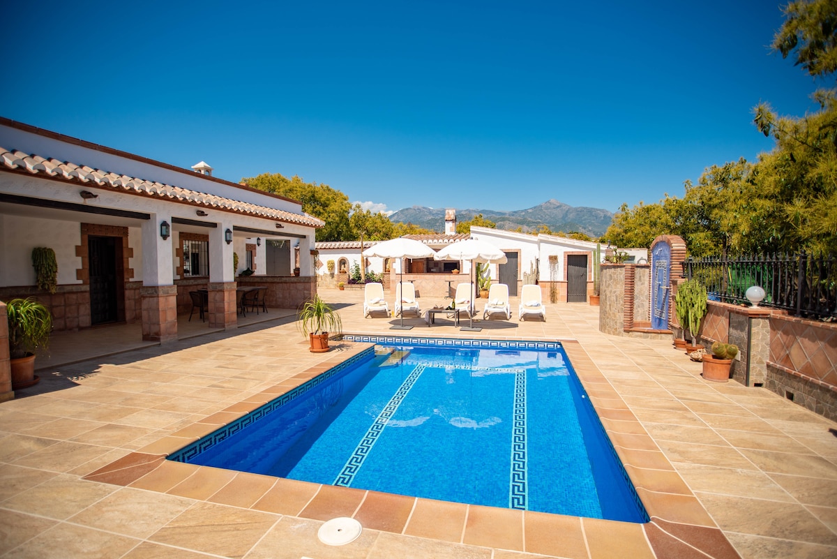 A private outdoor area is highlighted by a clear blue swimming pool surrounded by sun loungers. Umbrellas provide shade, while potted plants add a touch of greenery. The space is bordered by rustic architecture, and distant mountains can be seen under a clear blue sky.