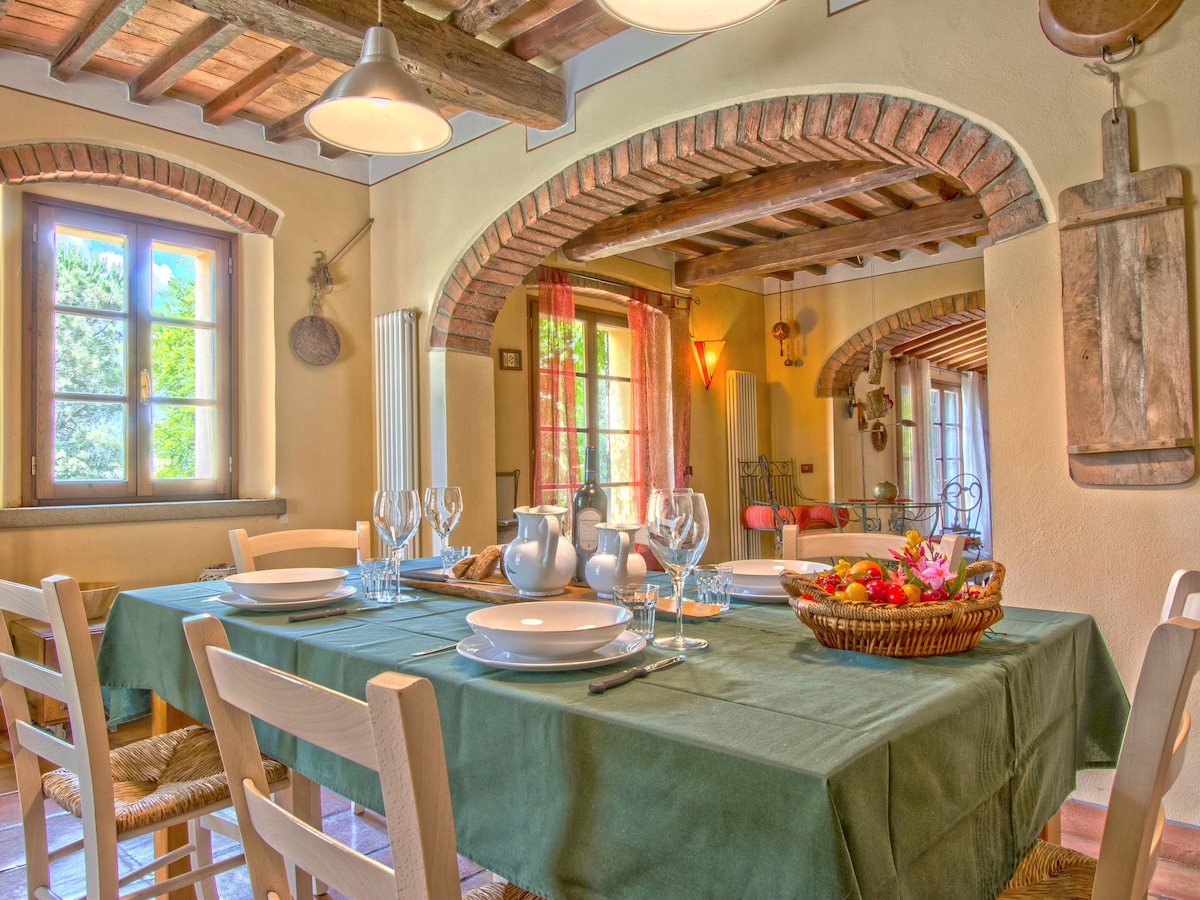 A welcoming dining area is set for a meal, featuring a large table with green cloth, dining ware, and a fruit basket. Natural light floods in through multiple windows framed by archways, highlighting rustic elements like wooden beams and terracotta tiles.