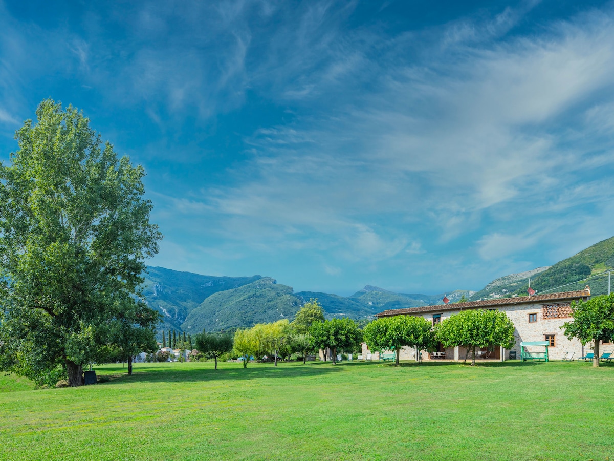 The image shows a spacious green lawn bordered by trees, leading to a semi-detached house with a rustic facade. Surrounding mountains create a serene backdrop under a clear, blue sky, suggesting a peaceful natural environment.