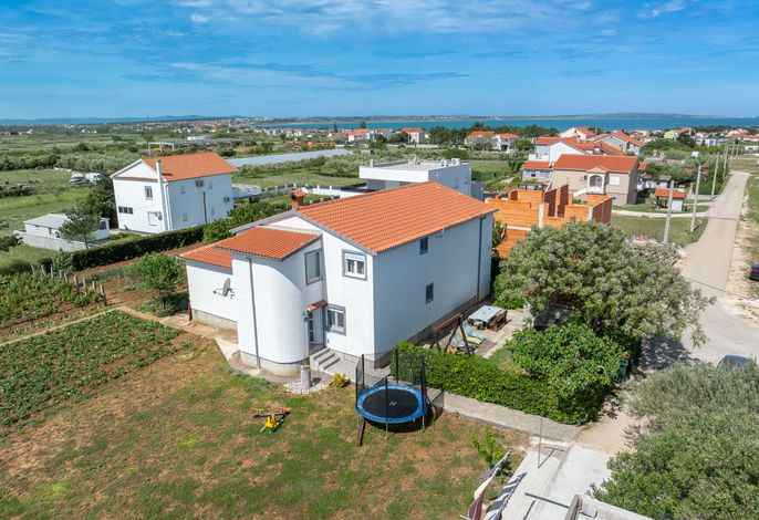 A two-story white building with a terracotta roof is visible. The property is surrounded by greenery, with its garden featuring a trampoline and outdoor seating. A clear view of the nearby coastline and blue skies can be seen in the background, enhancing the peaceful setting.