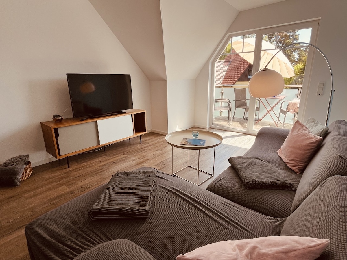 A spacious living area features a comfortable sectional sofa adorned with soft pillows. A coffee table sits in front of the sofa, and a large flat-screen TV is mounted on the wall. Sunlight streams in through the balcony doors, which lead to an outdoor seating area.