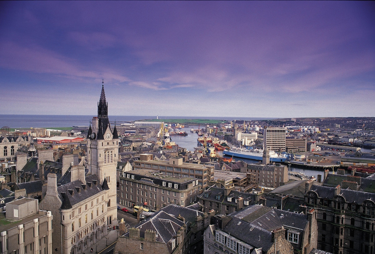 A panoramic view captures the bustling Aberdeen harbour, with vibrant boats docked in the water. The city's classic granite buildings are visible in the foreground, underscored by a dramatic sky transitioning from purple to blue, influencing the coastal atmosphere.