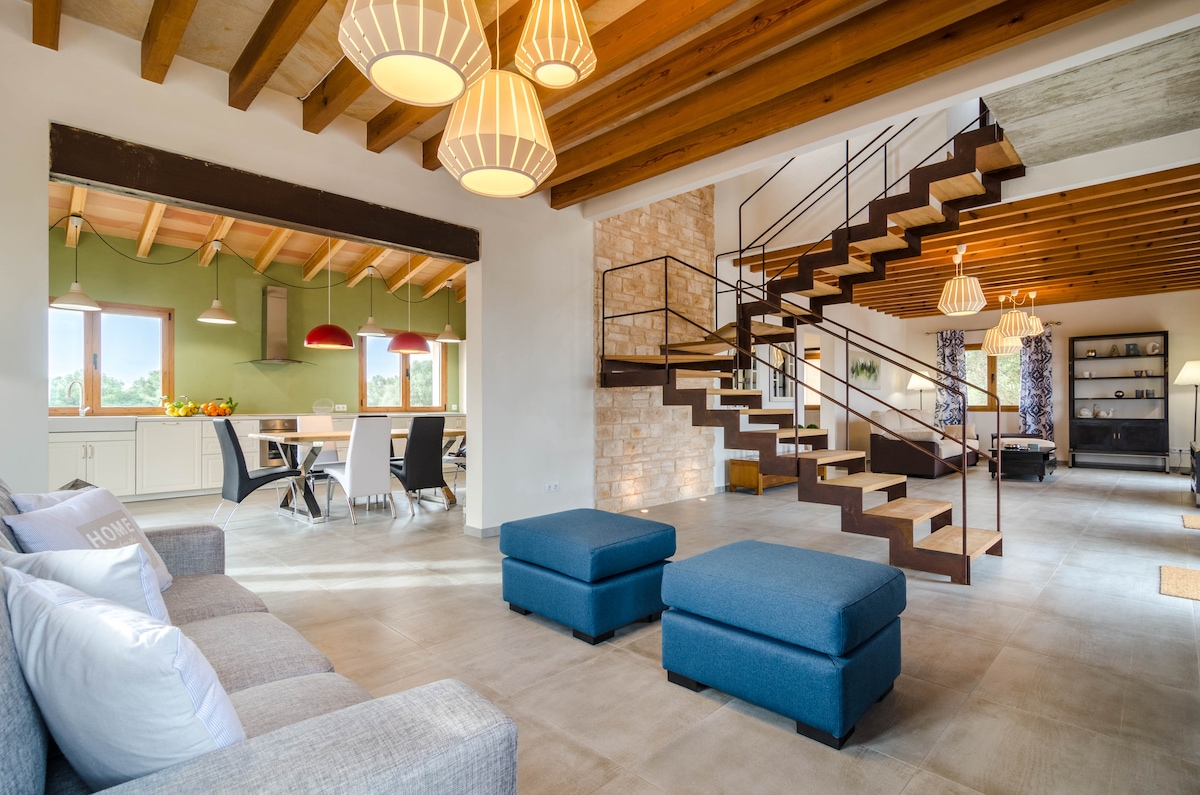 The living area presents a modern design with wooden beams and large windows that allow natural light to fill the space. A comfortable sectional sofa is positioned alongside two ottomans. A dining area is visible, equipped with a table and stylish chairs, enhancing the open layout.