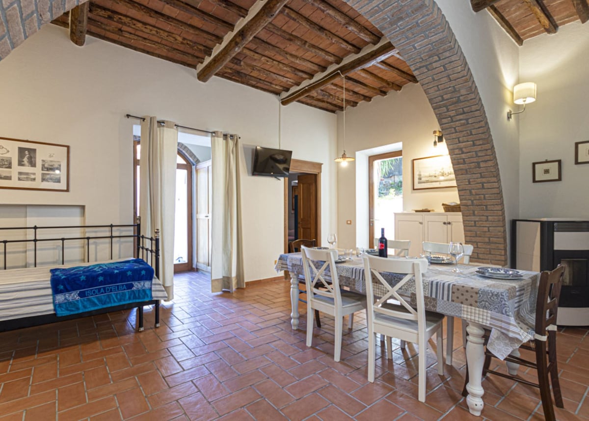 A spacious living area features rustic wooden beams and terracotta floor tiles. A dining table is set for six, complemented by a television mounted on the wall. A single bed is situated in the corner, adjacent to large windows that allow natural light to fill the room.