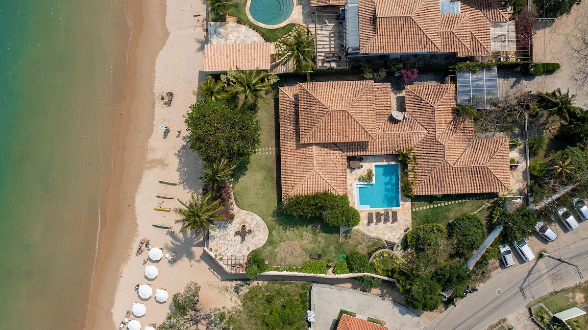 An aerial view showcases the property situated by the beach, featuring a swimming pool and a garden area with lounge chairs. The layout includes a sandy beach and well-maintained pathways lined with palm trees, emphasizing the spacious outdoor living spaces.