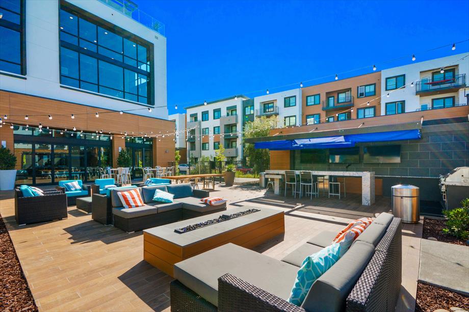 An outdoor courtyard features modern lounge seating with colorful cushions and several dining tables. A bar area with high-top seating is visible, and string lights are hung overhead, providing a relaxed atmosphere. The space invites communal gatherings and leisurely afternoons.