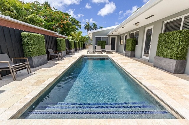 A rectangular pool is centrally located, surrounded by elegant lounge chairs and potted greenery. The pool's clear water reflects the blue sky and white clouds above, offering a serene setting amidst the sleek modern architecture of the building.