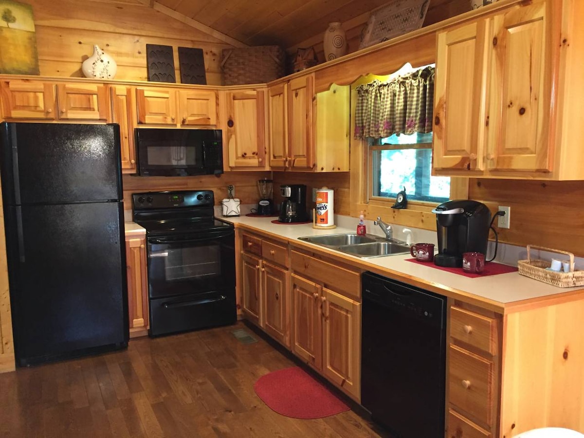 A warm wooden kitchen features ample cabinetry and modern appliances. A black refrigerator and microwave, along with a stove, are neatly arranged. Counter space is provided for food preparation, with a sink and a coffee maker ready for use. Natural light filters through a window adorned with a checkered curtain.