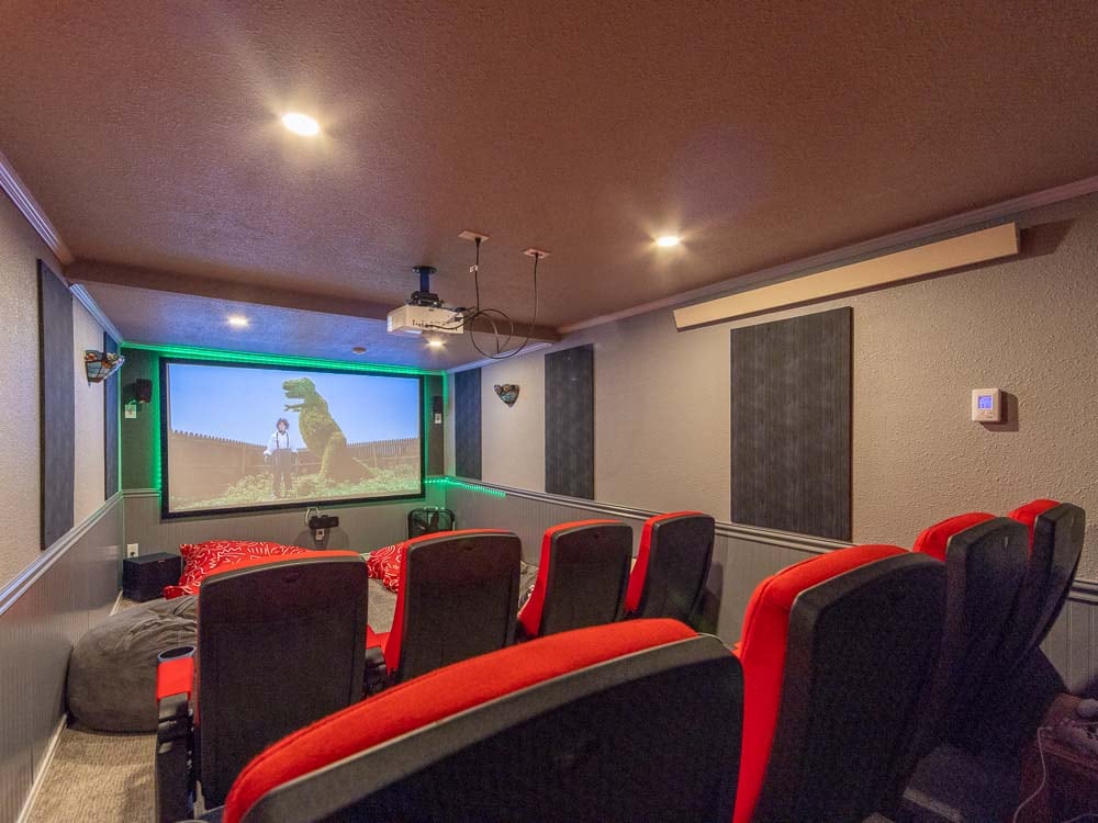 A private theater room is featured, equipped with plush red seating arranged in a tiered layout. A large screen at the front displays a movie. The walls are accented with black panels, and subtle green lighting enhances the viewing experience.