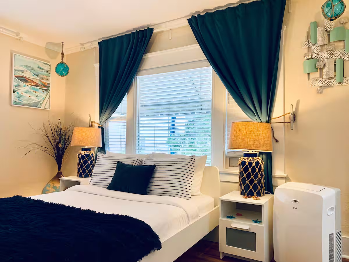A cozy bedroom features a spacious bed adorned with crisp white linens and a textured black throw blanket. Two bedside lamps provide warm lighting beside the bed, framed by flowing teal curtains. A decorative wall art piece adds color above the headboard, complemented by an air conditioning unit.