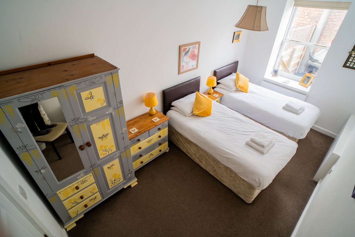 A well-lit bedroom featuring two spacious beds with crisp white linens. Each bed is adorned with golden-yellow cushions. A colorful wardrobe and matching bedside tables are present, alongside a reflective mirror. Natural light enters through a window, creating a bright and inviting environment.