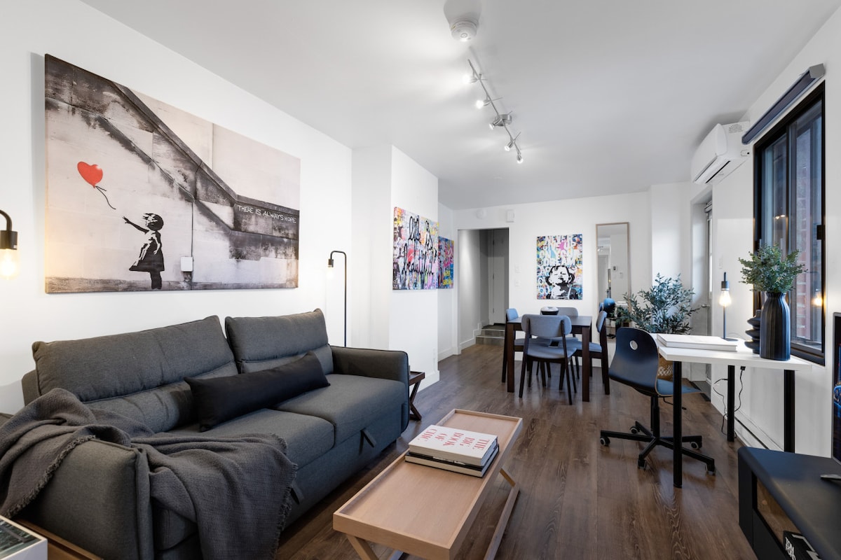 The spacious living area features modern furnishings, including a comfortable sofa with a cozy throw. Colorful curated artwork adorns the walls, while a dining table surrounded by chairs is positioned near the natural light from large windows.