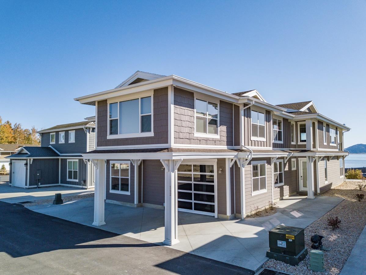 The exterior of a multi-level lakefront home showcases a modern architectural design with a gray color palette. Large windows and a two-car garage provide ample light and convenience. A clear blue sky and the nearby lake create a serene backdrop.