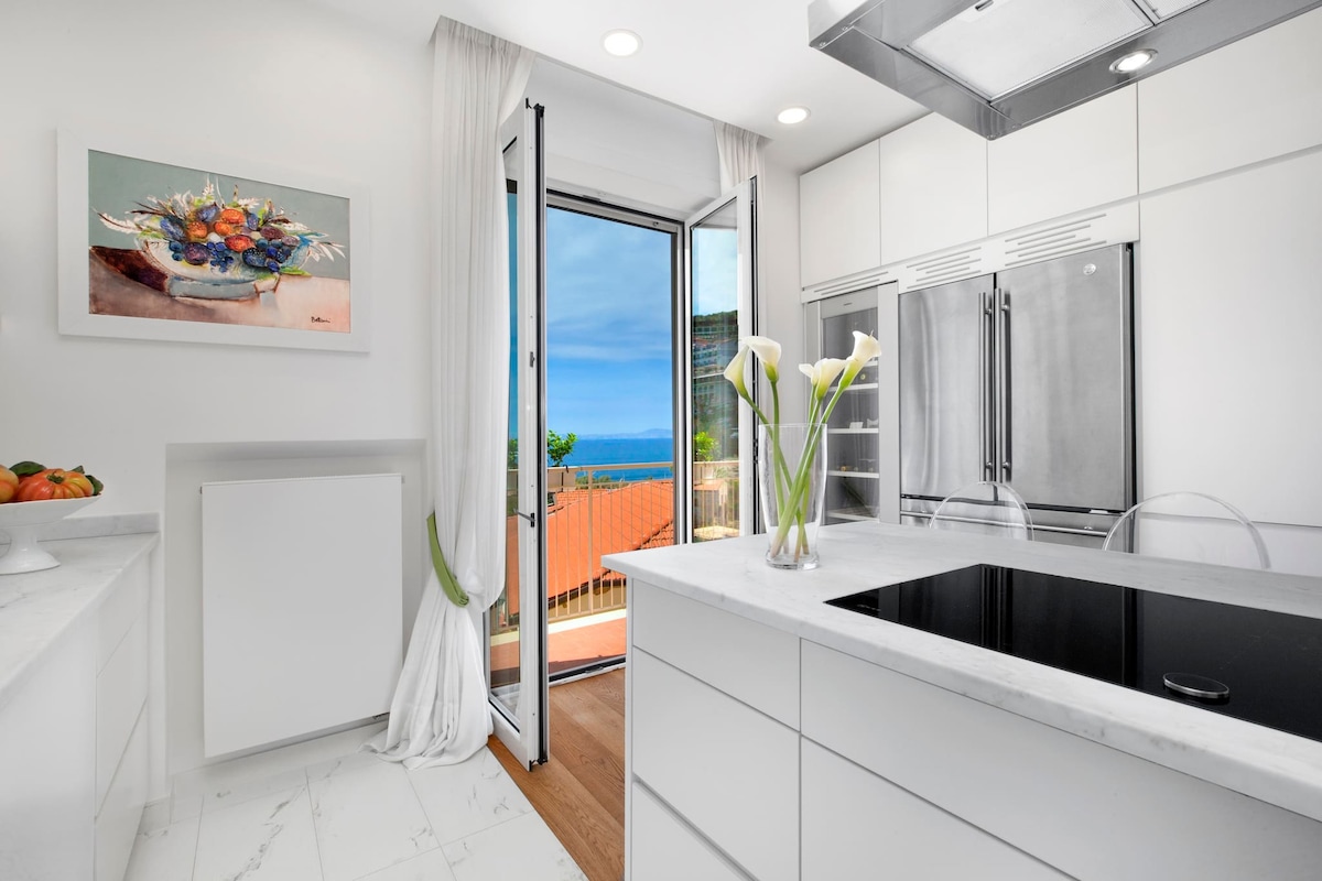 The modern kitchen features a Carrara marble island with transparent chairs, complemented by stainless steel appliances. Fresh flowers are displayed on a countertop. Open doors reveal a balcony with a sea view, allowing natural light to brighten the space.