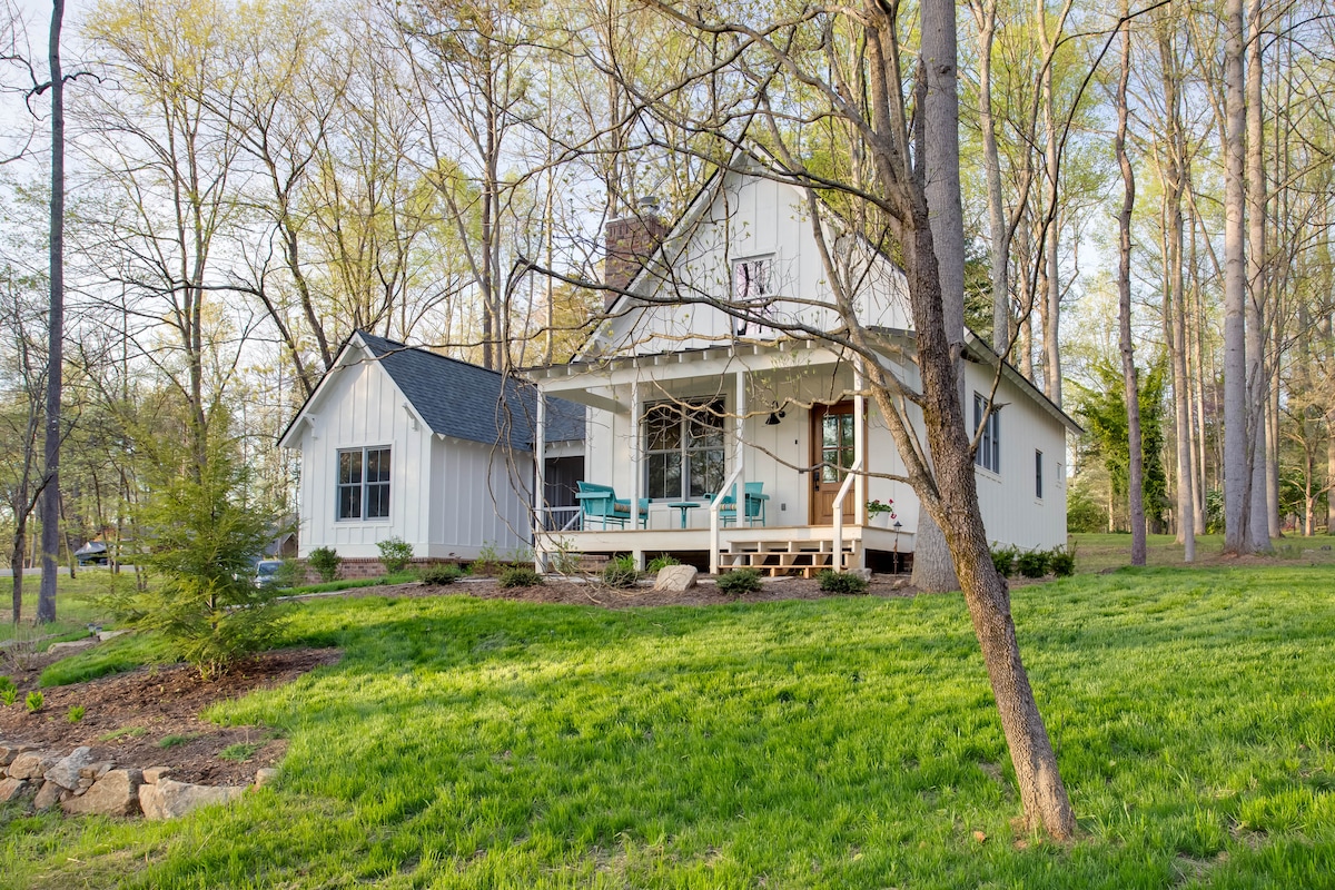 A charming two-story home is nestled among trees, featuring a welcoming porch with teal seating. The exterior displays a combination of light-colored siding and a contrasting dark roof. Lush greenery surrounds the property, enhancing the tranquil atmosphere of the setting.