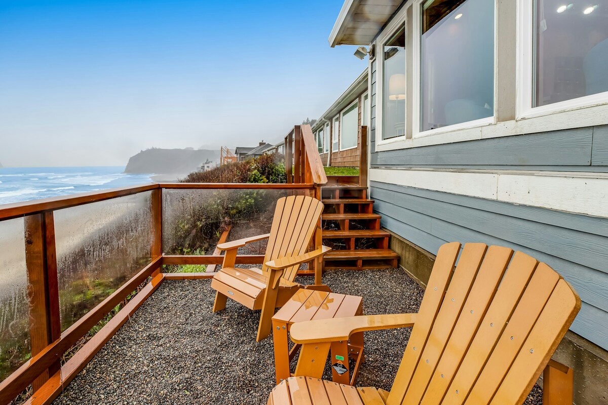 Two wooden Adirondack chairs are positioned on a deck overlooking the ocean, offering scenic views of the beach and waves. The deck is surrounded by a glass railing, providing unobstructed sightlines to the ocean, while steps lead down to the sandy beach below.