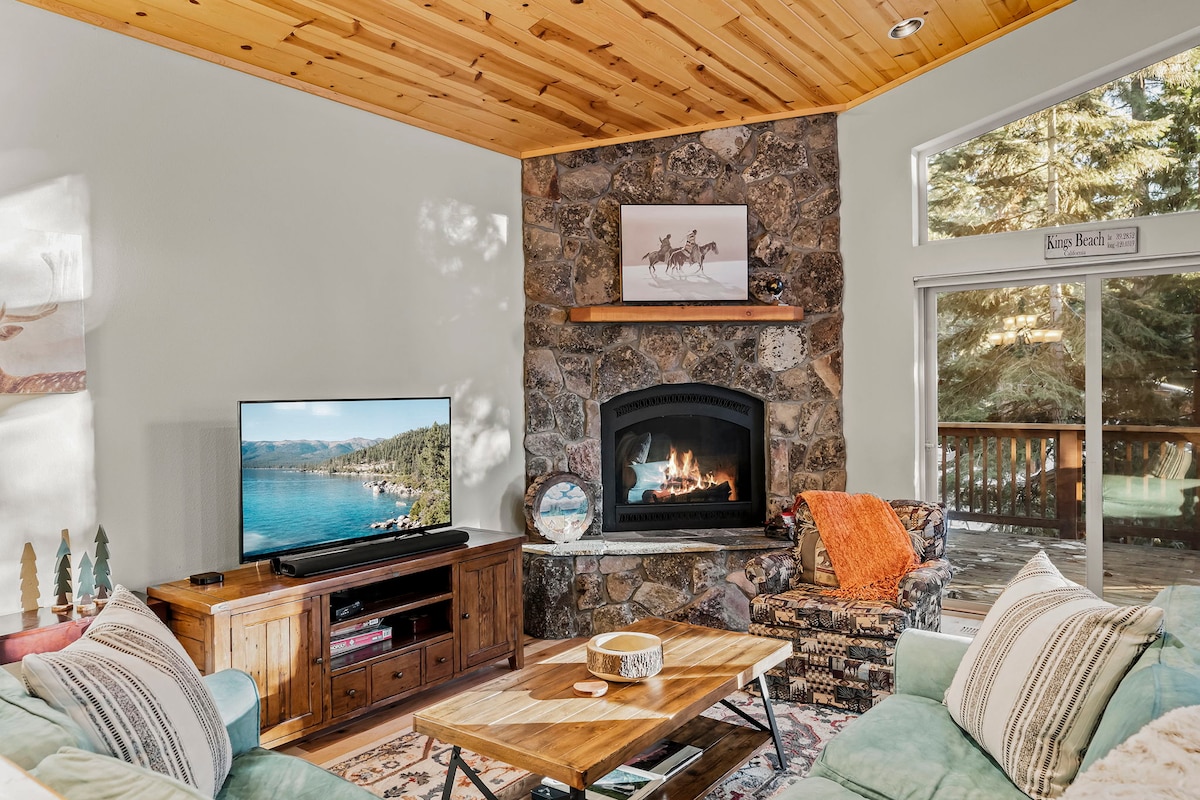 A cozy living area features a stone fireplace, complemented by a wooden ceiling and wall-mounted television. The space includes a comfortable sofa with colorful throw pillows and a wooden coffee table. A large window invites natural light and offers a view of the outdoor deck.