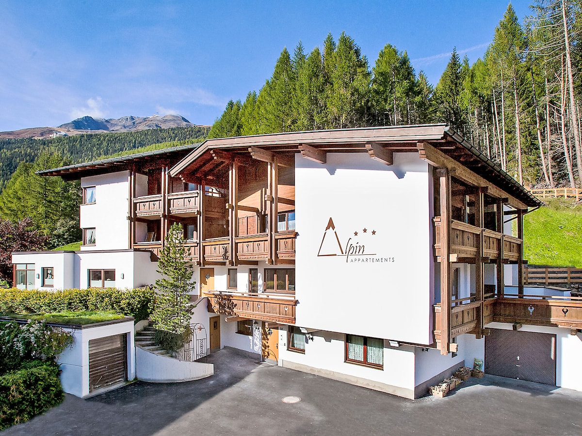 The exterior of the Alpin apartment building is showcased, featuring a mix of traditional and modern architectural elements. Surrounding trees frame the property, while the mountains are visible in the background, indicating a tranquil setting. Ample outdoor parking is provided in front.
