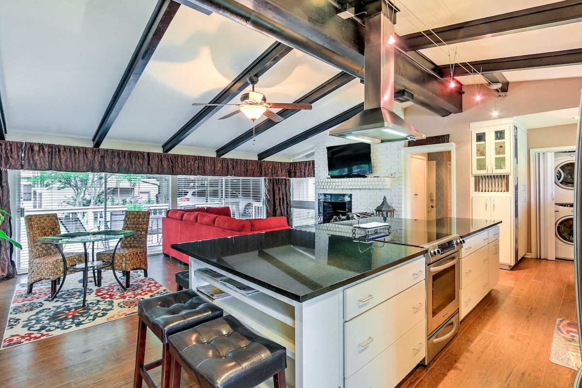 An open-concept kitchen and living area is highlighted by high, beamed ceilings. A spacious island with a dark countertop offers seating. The living space features a bright red sectional sofa, complemented by a coffee table and a television mounted above a cozy gas fireplace.