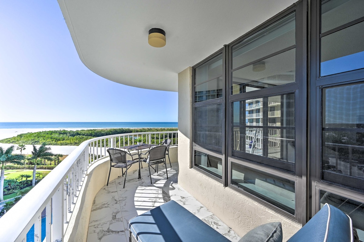 A spacious private balcony is shown, furnished with two comfortable chairs and a chaise lounge. The view showcases the Gulf of Mexico and a lush nature preserve, with bright sunlight filling the area through large windows.