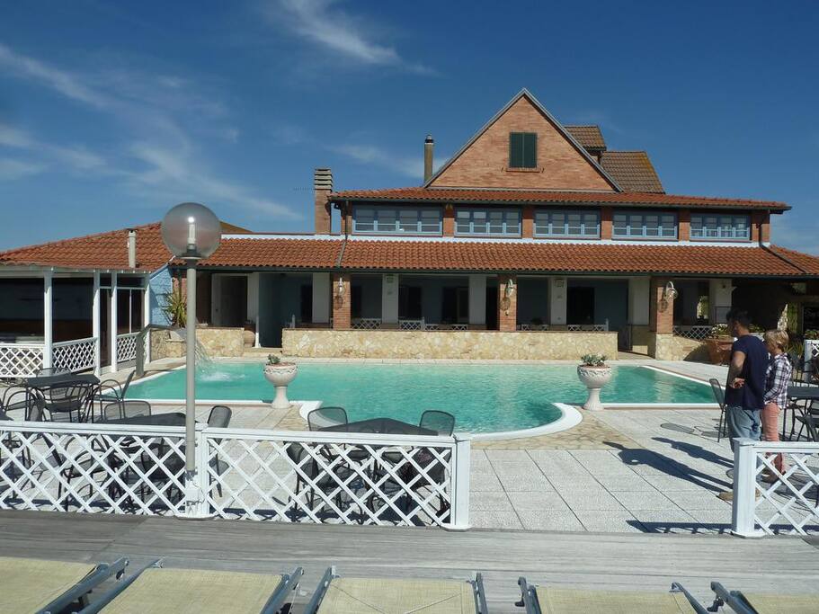 The exterior of a large building is visible, featuring a terracotta roof and expansive glass windows. A rectangular swimming pool is situated at the center, surrounded by a spacious deck with seating areas. Lush greenery adds to the inviting atmosphere.
