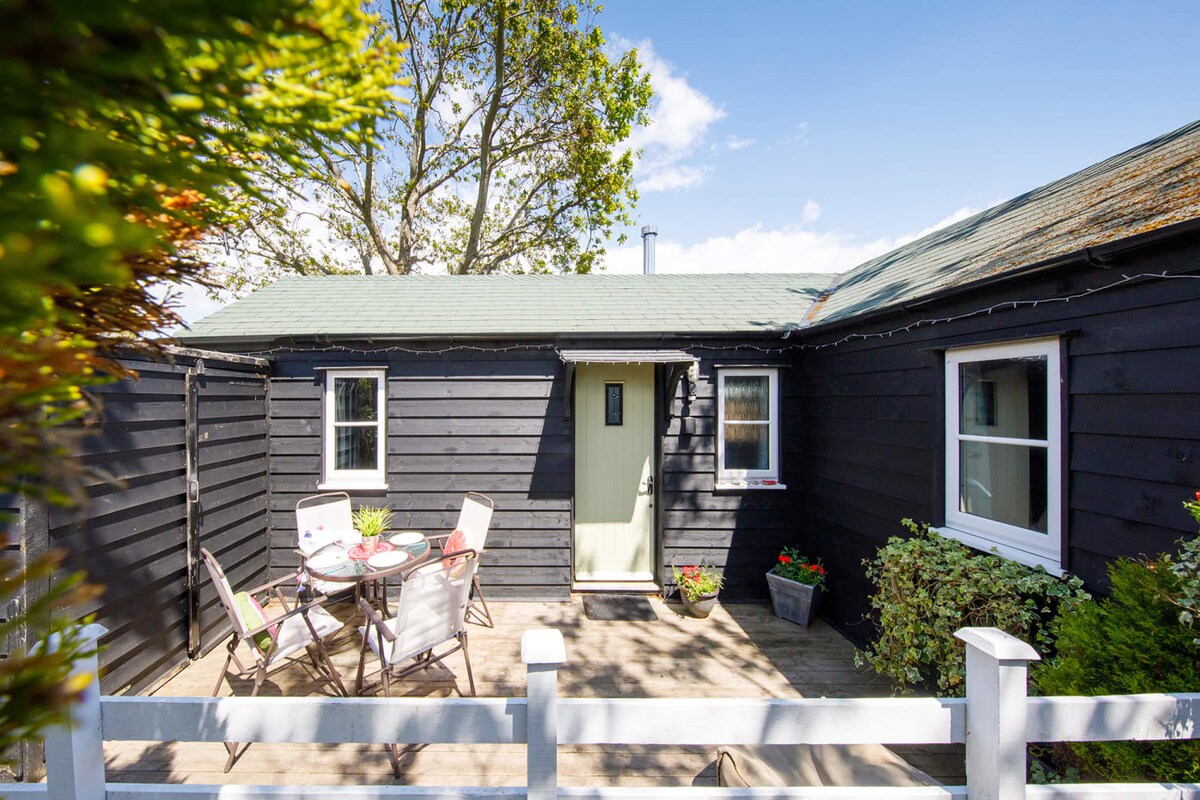 The exterior of the annexe is displayed, showcasing a wooden structure with a green roof. A small deck area with a table and chairs is visible, surrounded by potted plants and greenery. Two windows provide natural light, and a private entrance is seen.