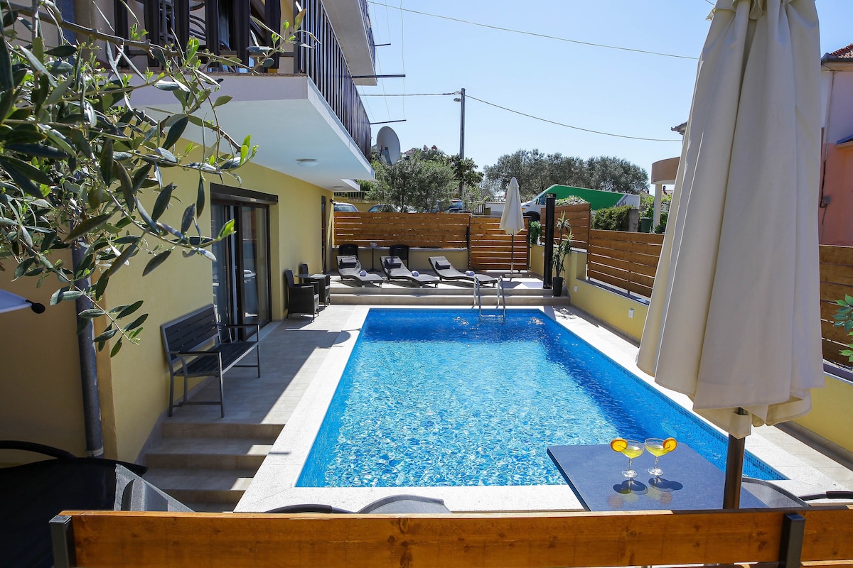 The shared pool area features a clear blue swimming pool surrounded by a stone deck. Sun loungers are positioned nearby, and shaded seating is provided to one side, offering a space to relax. A parasol stands by the poolside, enhancing the usability of the area.