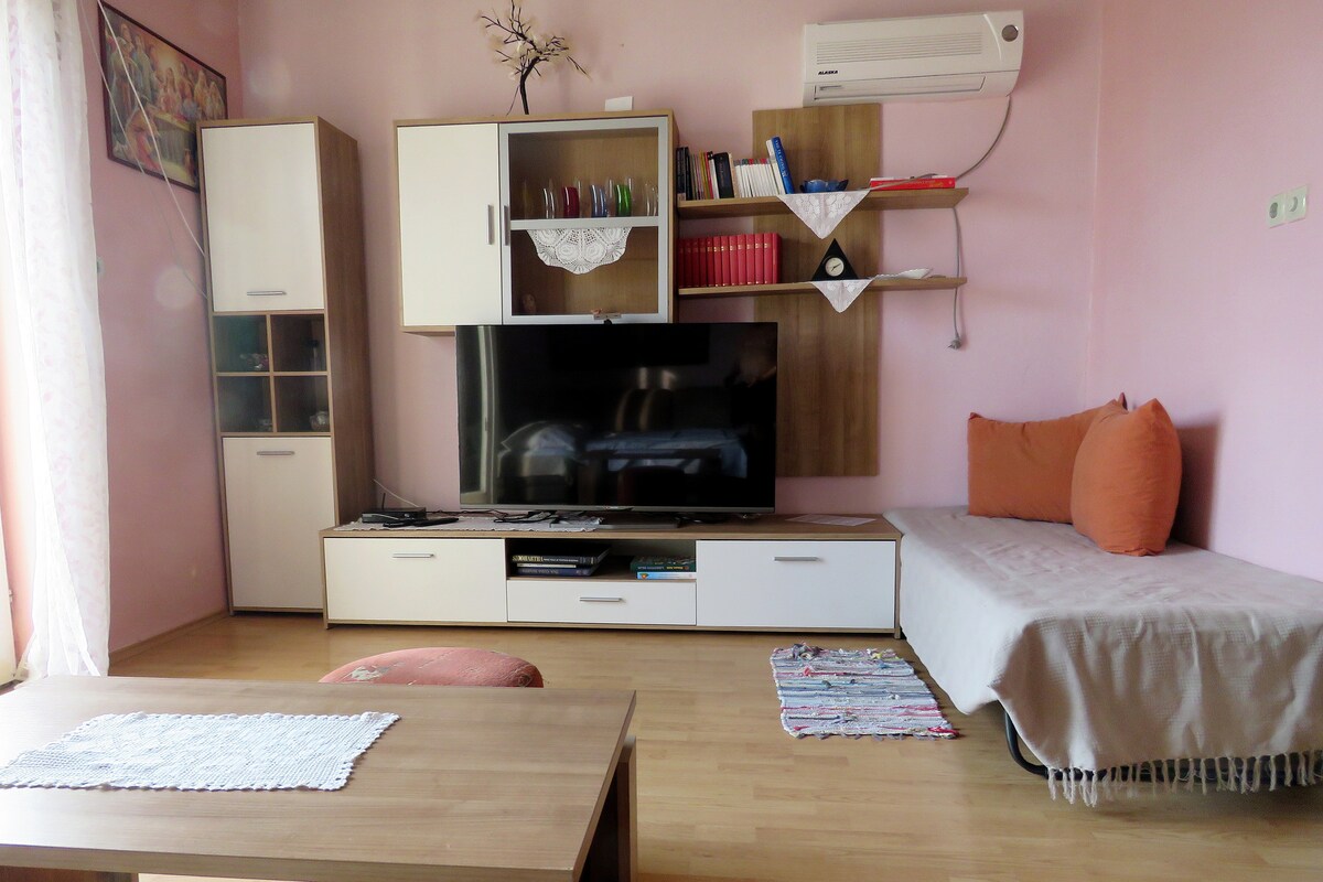 A living area is furnished with a wall unit that includes a television and shelving. A comfortable seating area is present, featuring a spacious sofa adorned with orange cushions. Natural light floods the room through partially draped windows, with light-colored walls enhancing the space's brightness.