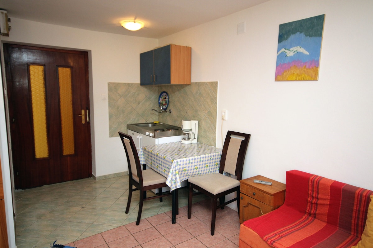 A studio flat features a dining area with a white table covered by a checkered tablecloth and four chairs. A small kitchen is visible, along with a red sofa placed against the wall. Light walls and an art piece above the kitchen enhance the minimalistic design.