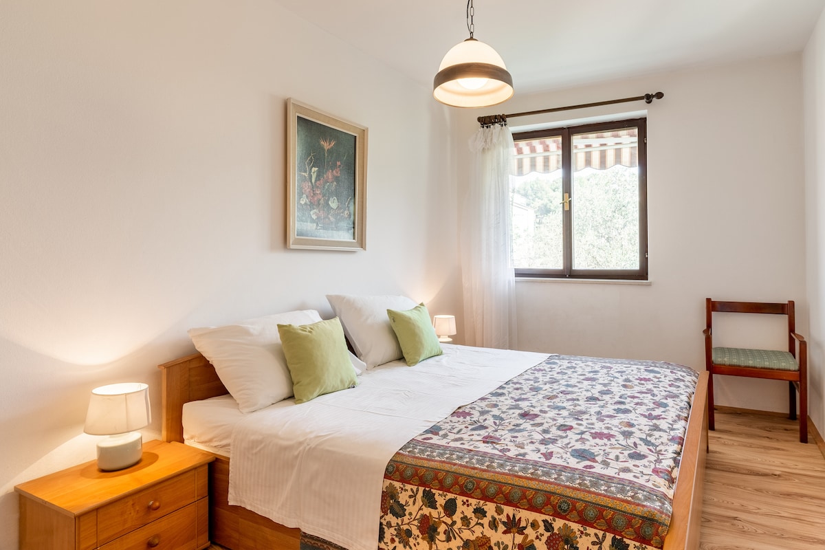 A bright bedroom features a spacious bed with light linens and decorative pillows. A colorful textile runs along the edge of the bed. Natural light enters through a window, with a simple chair placed in one corner. The walls are painted in a soft white.