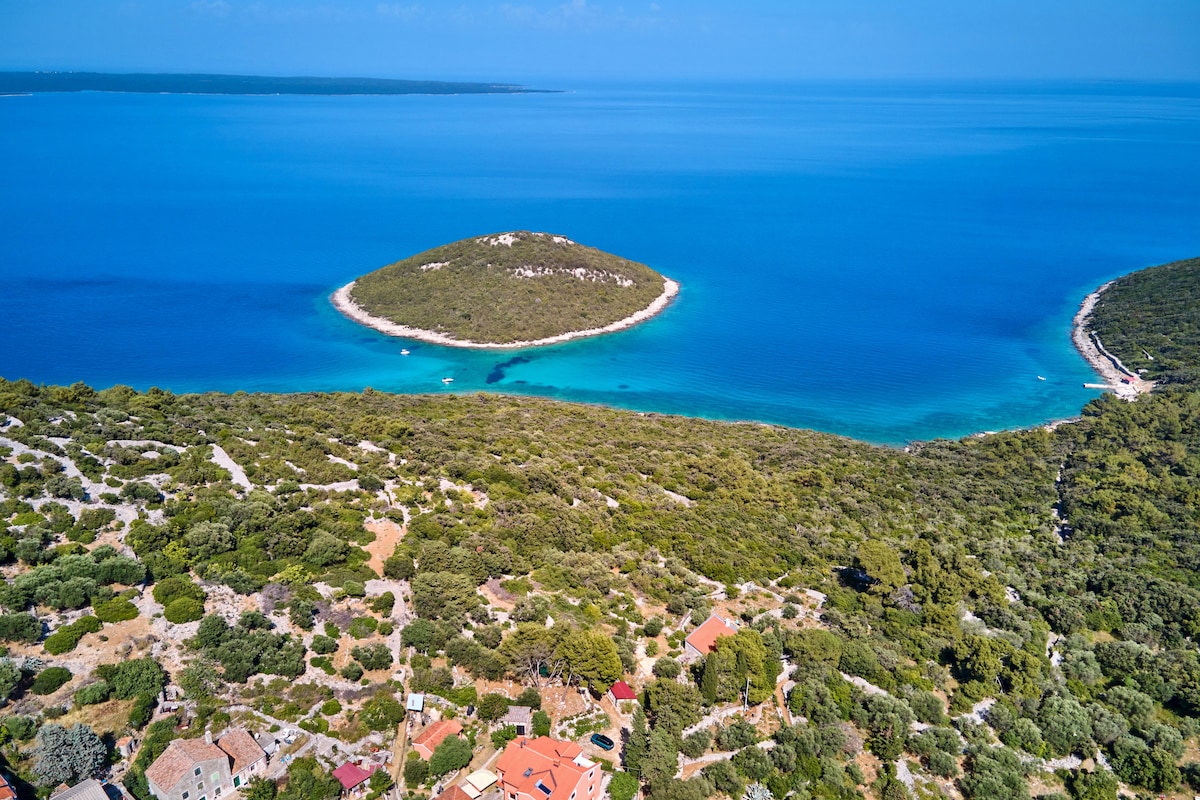 An aerial view captures the serene landscape of the coastline, where turquoise waters meet an island surrounded by lush greenery. The clear sea contrasts with the rocky shorelines, inviting exploration of the natural beauty of the area.