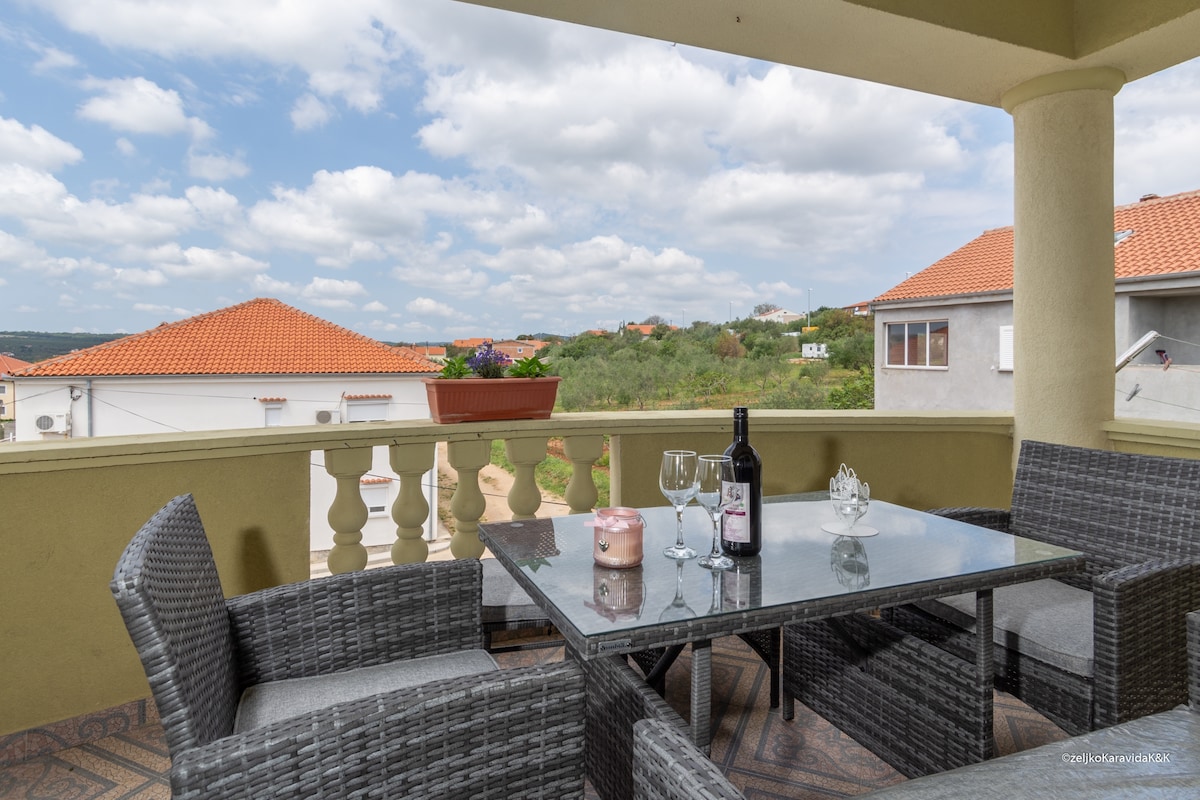 A private terrace features a glass-topped table surrounded by four wicker chairs. Two wine glasses and a bottle of wine are displayed on the table. The view includes distant rooftops and greenery, complemented by a cloudy sky.