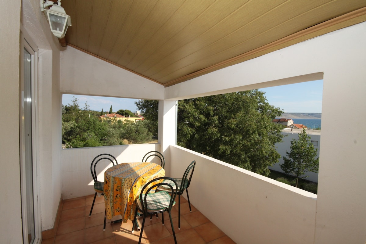 A private terrace is shown, featuring a round table covered with a patterned tablecloth and four metal chairs. Large openings provide views of surrounding trees and the distant sea, enhancing the outdoor experience.
