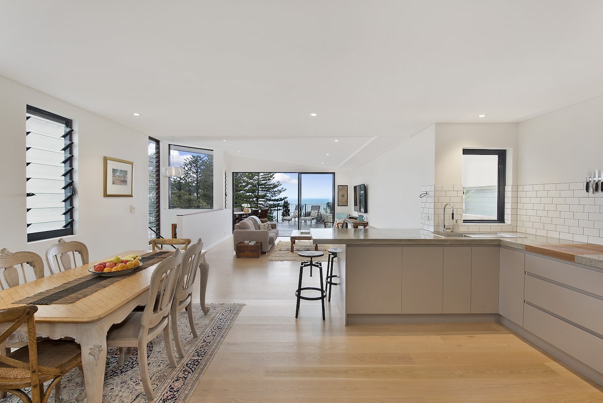 An open-plan living and dining area is showcased with a large wooden table set for dining. A modern kitchen features sleek countertops and minimalist cabinetry. Floor-to-ceiling windows provide a view of the outdoors, enhancing natural light and connecting the interior with the scenery.