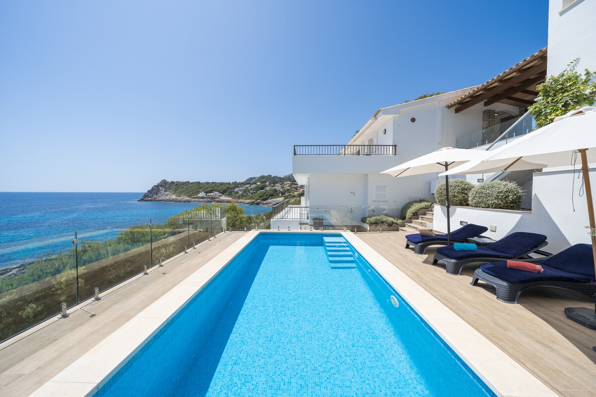 A clear blue swimming pool is surrounded by sun loungers, positioned next to a glass railing that offers views of the ocean. The terrace features umbrellas providing shade, with the coastline visible in the background under a bright sky.