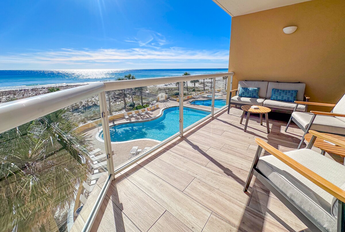 The Gulf-front balcony is furnished with two chairs and a small table, offering views of the beach and shimmering sea. The outdoor space overlooks a pool surrounded by sun loungers and lush greenery, providing a relaxing retreat in a well-lit environment.