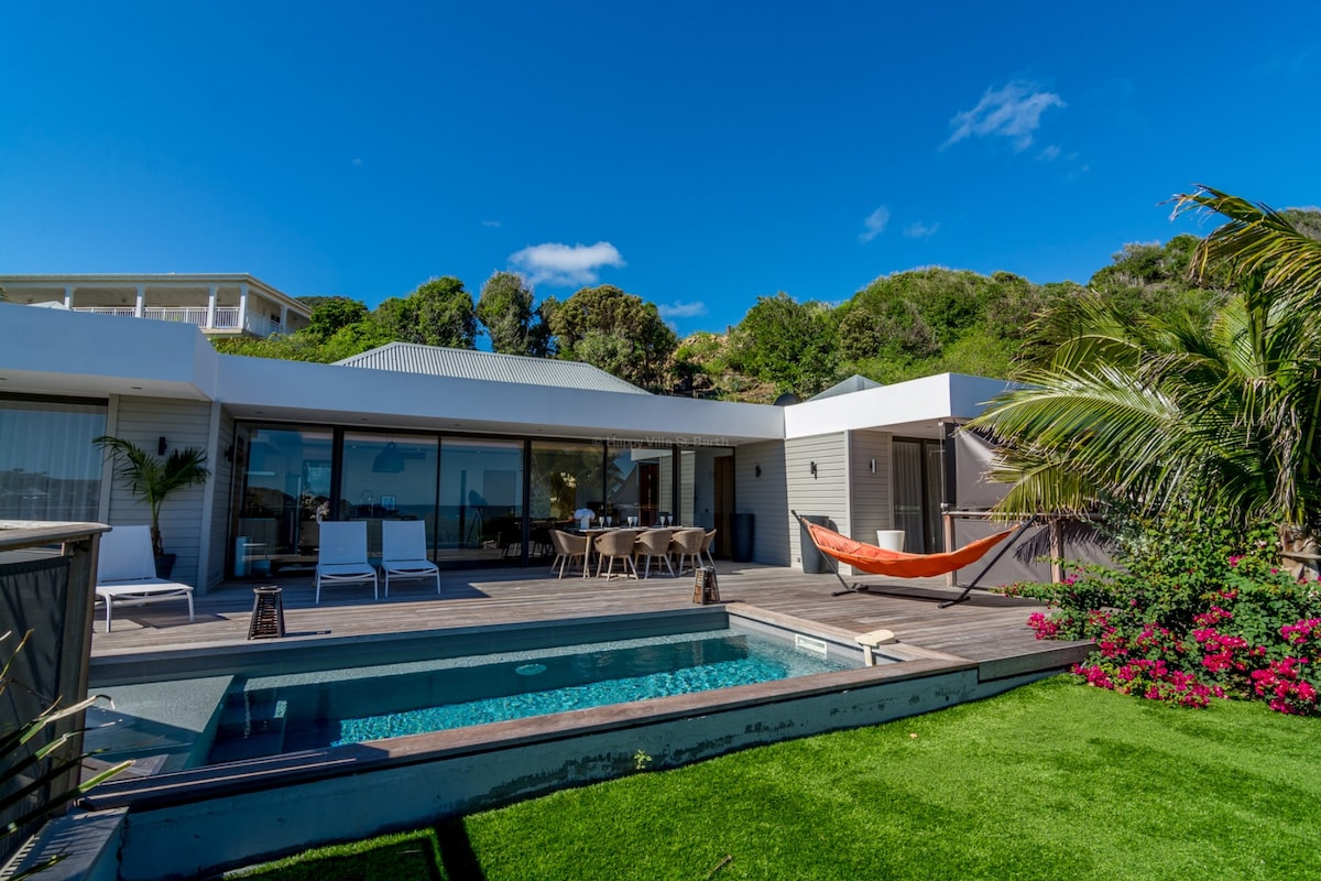 The exterior of the villa features a spacious deck area surrounding a heated pool. Comfortable seating options are available, including a dining table, lounge chairs, and a hammock. Lush greenery and colorful flowers frame the scene, enhancing the tranquil outdoor setting.
