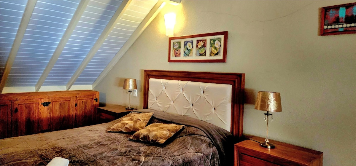 A comfortably arranged bedroom features a spacious bed with a plush blanket and decorative cushions. Bedside tables with lamps provide soft lighting. The room is complemented by a framed art piece above the headboard and angled ceiling beams, enhancing the cozy atmosphere.