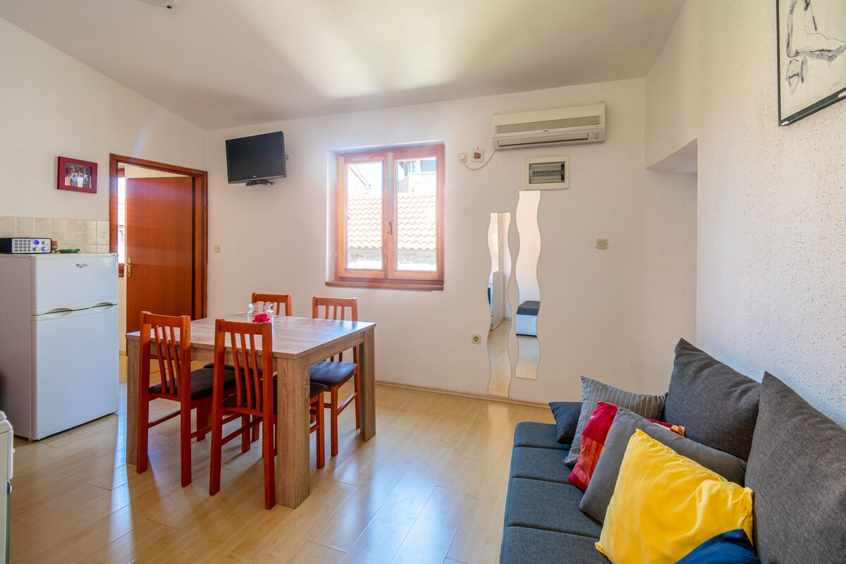A bright living area features a dining table with six wooden chairs and a comfortable sofa adorned with colorful cushions. Natural light enters through two windows, highlighting the air-conditioning unit on the wall. A small kitchenette is visible, equipped with essential appliances.